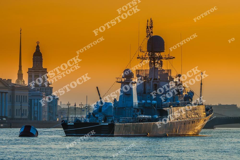 На фотографии изображен военный корабль, стоящий на Неве в Санкт-Петербурге во время заката. На фоне корабля видны знаменитые петербургские достопримечательности, такие как Кунсткамера и шпиль Петропавловского собора, добавляющие кадру историческую ценность. Оранжевое небо заката окрашивает воду и усиливает ощущение спокойствия и торжественности момента. Вечерняя подсветка придает снимку выразительность и особую атмосферу.